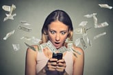 Shocked-looking woman staring at dollar bills flying out of her cellphone.