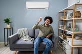 Young happy man sitting on couch operating air conditioner with remote control