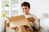 A happy man smiles while opening a package in a shipping box on his sofa