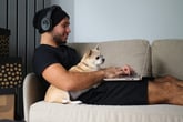 Smiling man laying on the couch with his dog while wearing headphones and shopping or playing a video game on his laptop.
