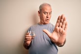 Older man holding up his hand in a stop gesture and holding a glass of water saying no