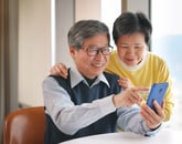 Senior couple smiling and looking happy while pointing at smartphone in front of a bright window at home