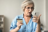 Surprised older woman holding her phone and credit card shopping online and surprised by a discount or fraud