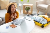 Excited woman with laptop and travel suitcase unpacked sitting in the living room with warm and bright light and a credit card with her passport and phone