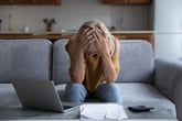 Stressed woman sitting on the couch holding her head in her hands feeling overwhelmed by financial paperwork or bills with a calculator and a laptop
