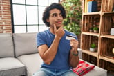 Thoughtful smiling man holding a credit card sitting on the couch at home doing online shopping or planning to use credit card rewards