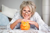 A senior woman in glasses smiles while he rests her hands on a piggy bank and lies on a bed
