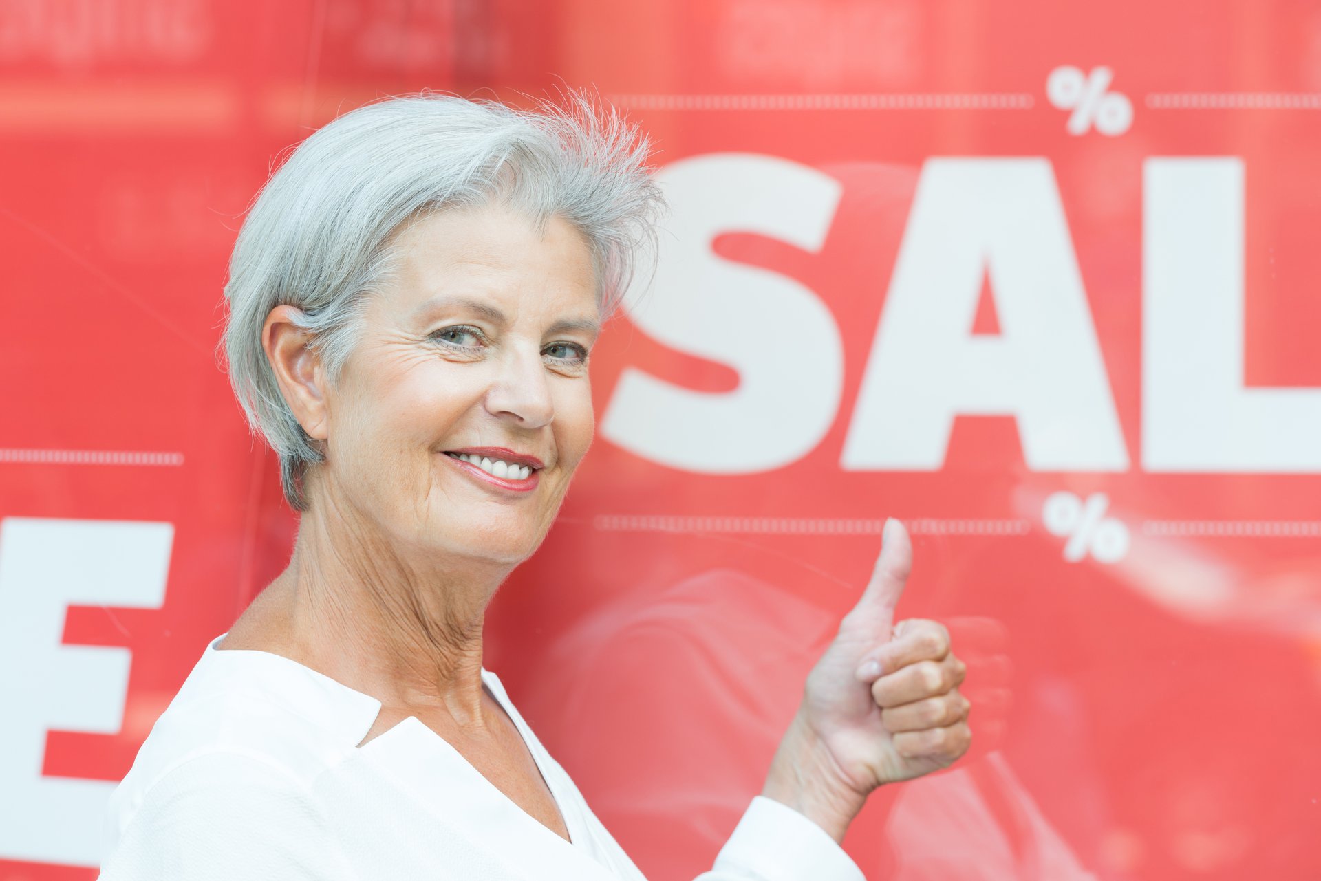 A senior woman gives a thumbs-up in front of a sale sign