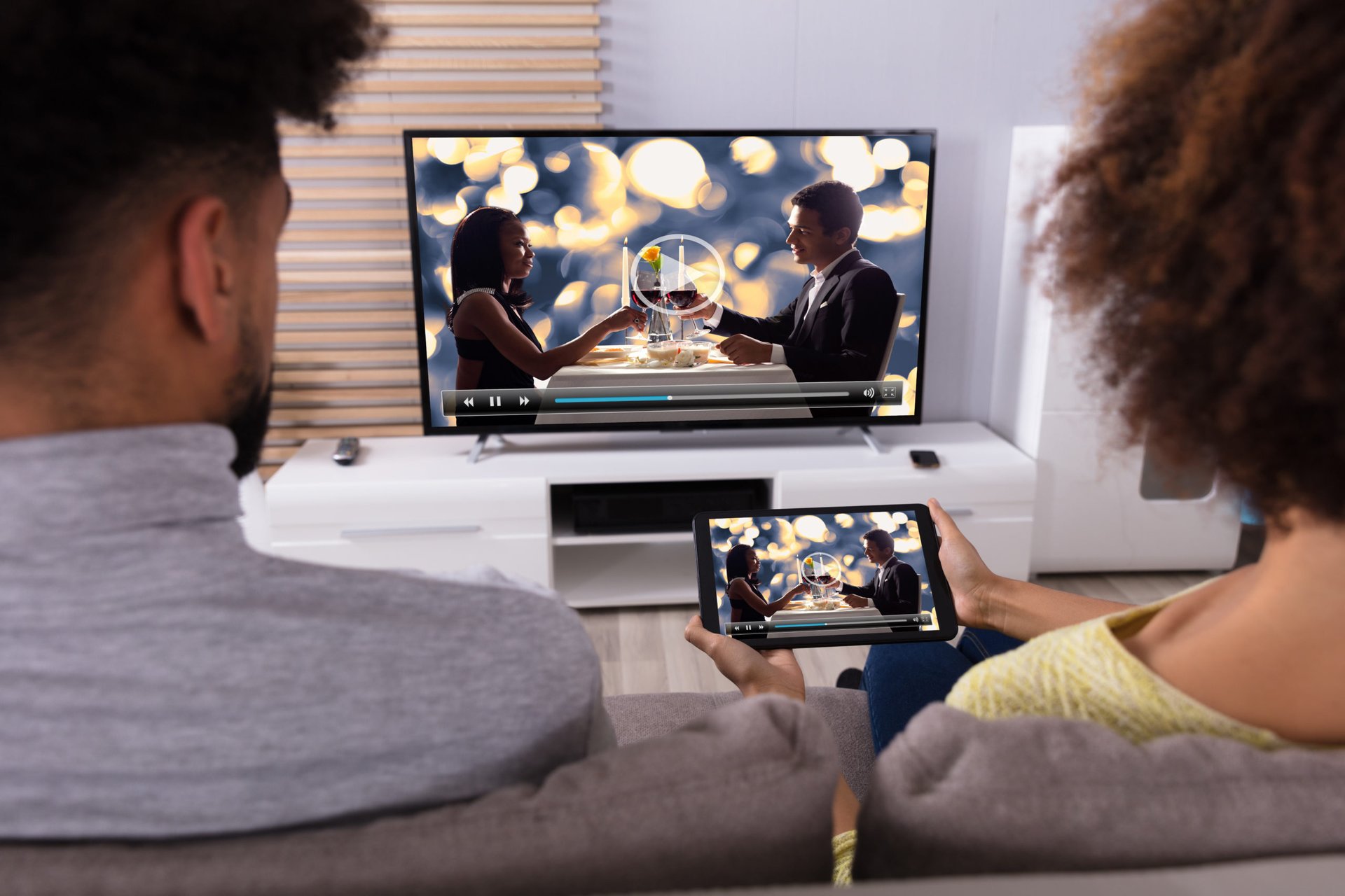 A couple watches TV on a TV and a tablet from their living room sofa