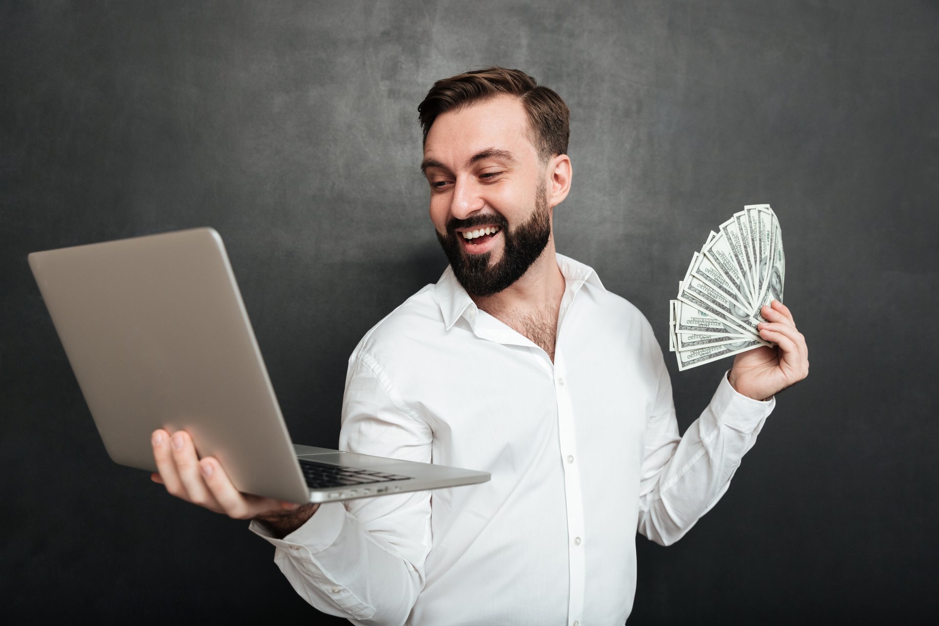 A happy man holds a laptop computer and fan of cash