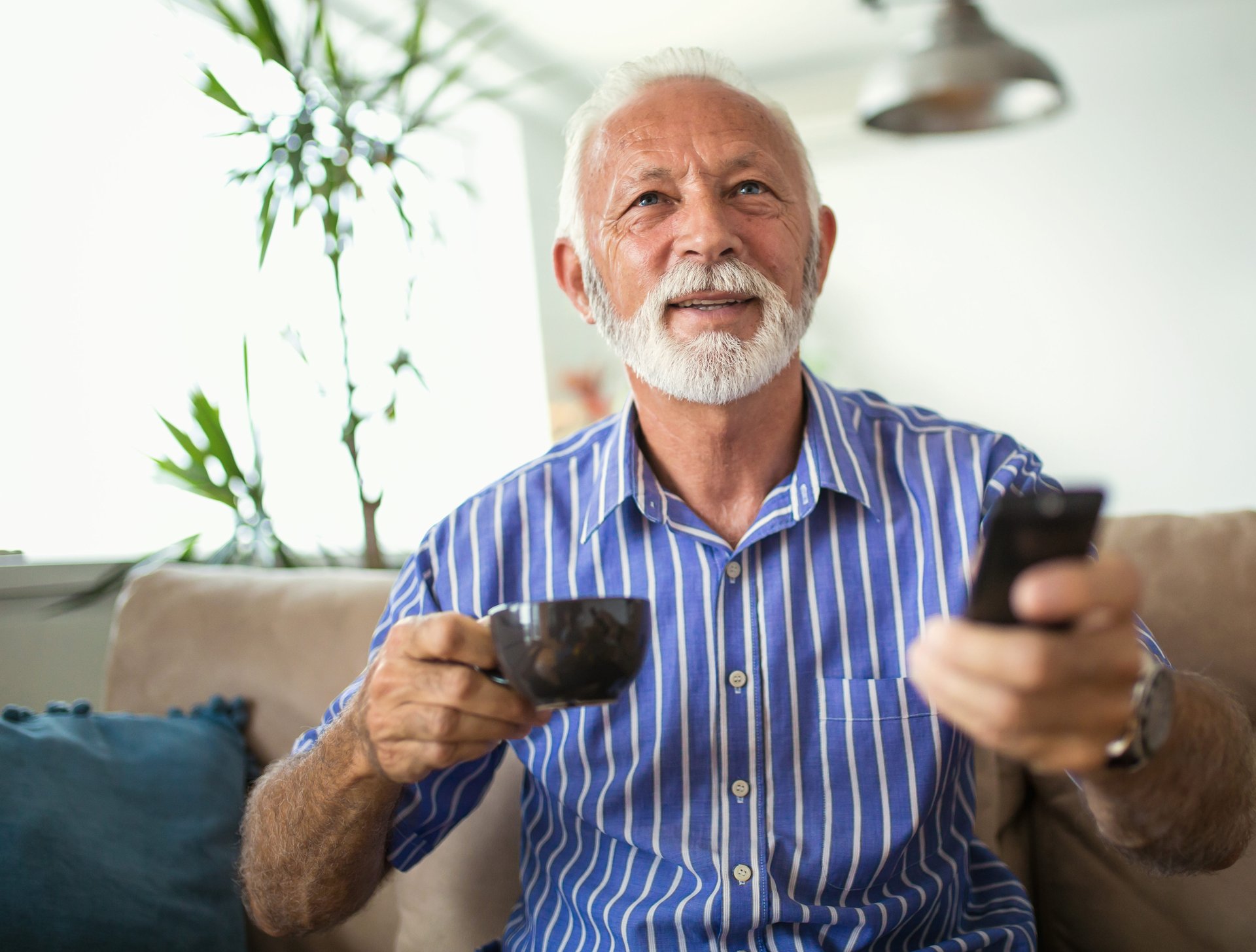 A senior man holds a remote control and coffee while watching tv on his sofa