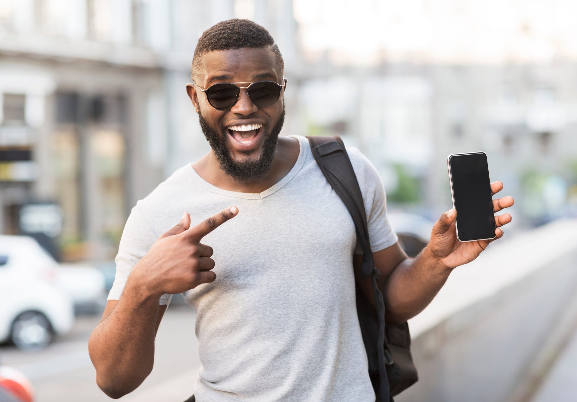 A Black man in sunglasses and a t-shirt excitedly points at his new cell phone