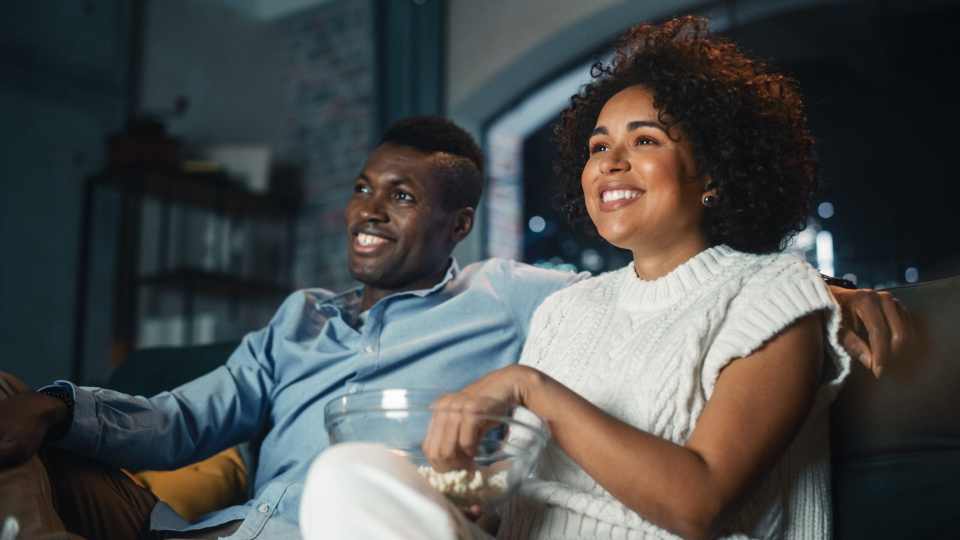 Smiling happy couple watching a movie or TV show on TV with home streaming service or cable while eating popcorn