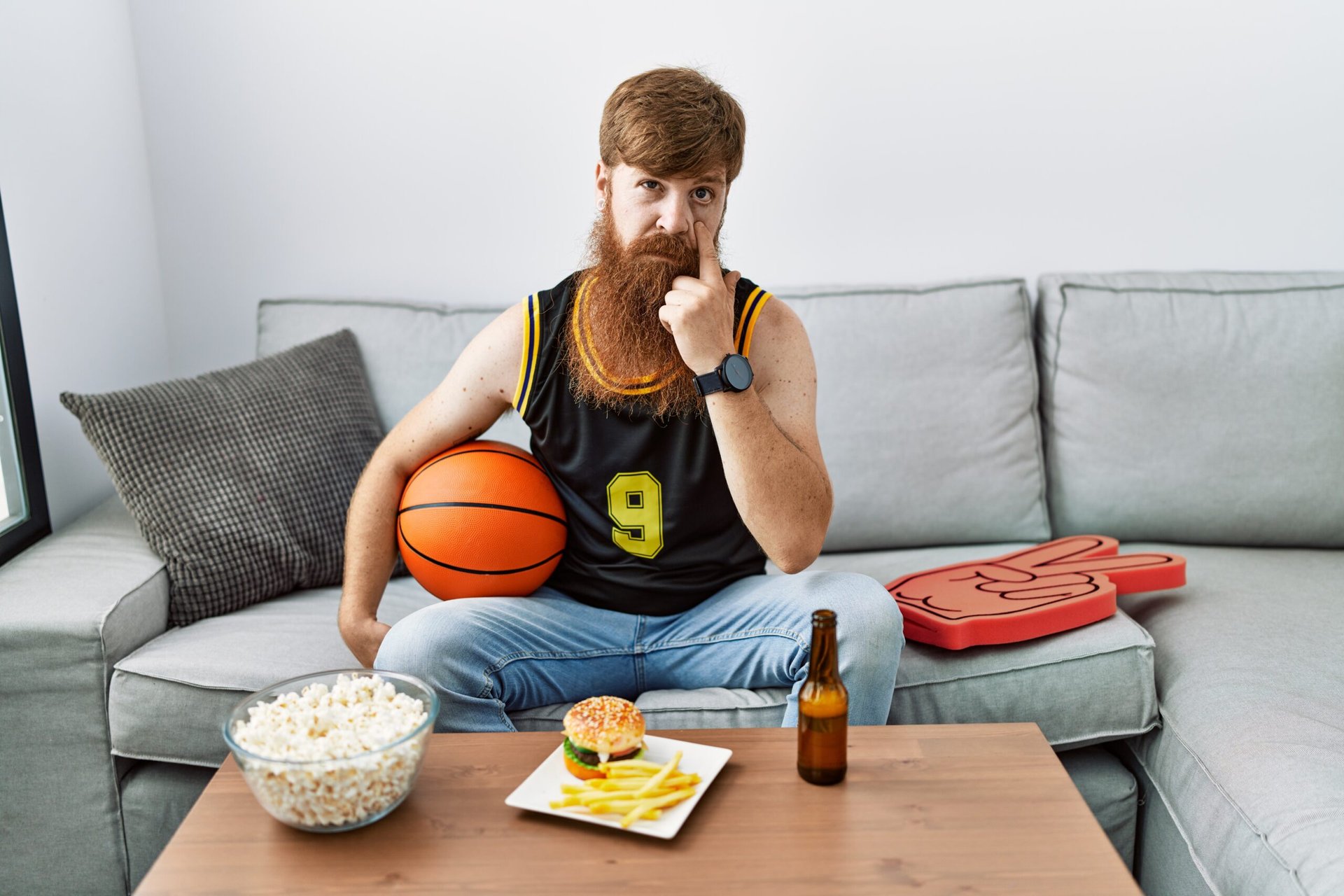 Bearded man holding a basketball watching TV with popcorn and a cheeseburger wearing a sports jersey and a watch sitting on a couch and making a watchful gesture