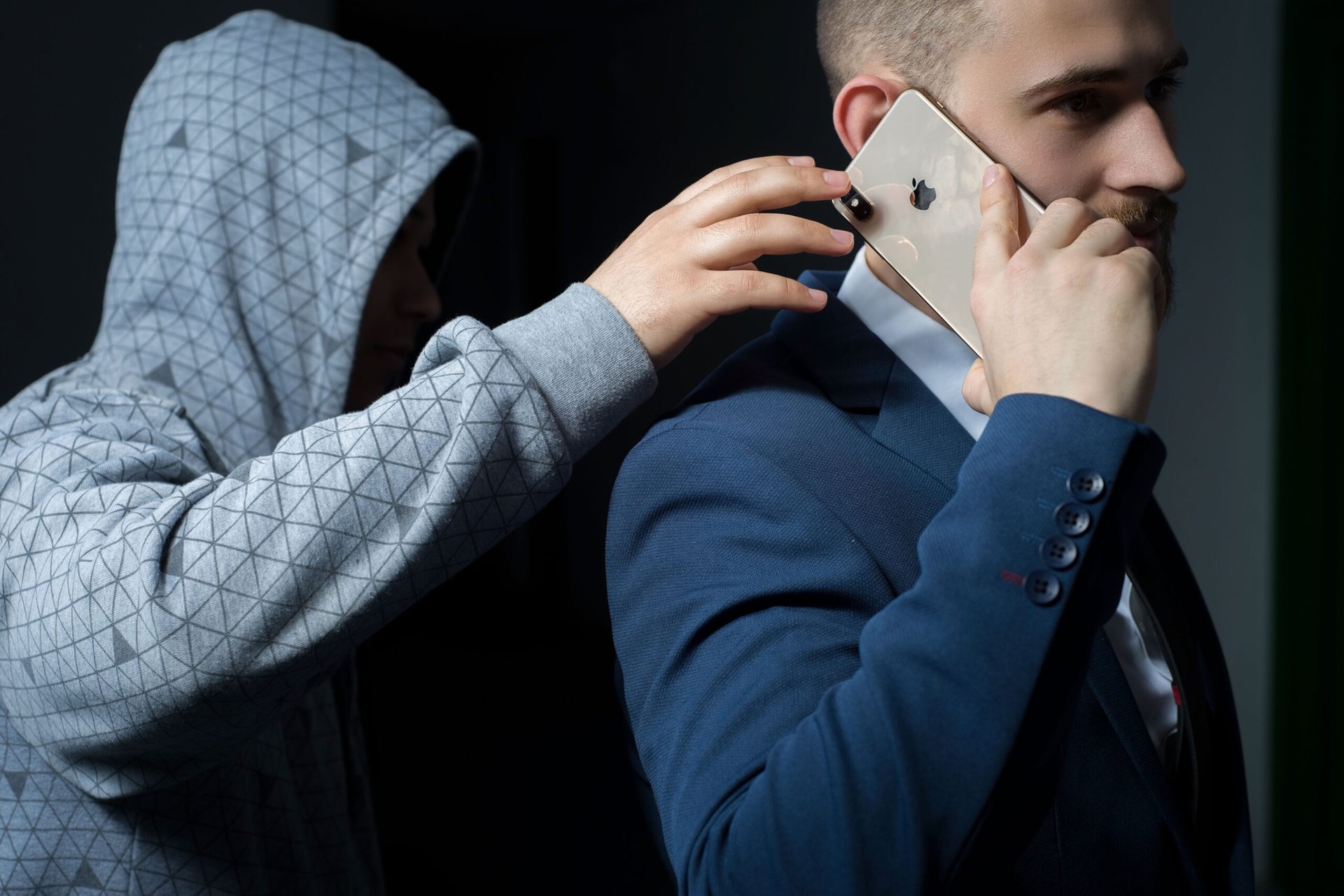 Thief wearing a hoodie stealing an iPhone from a businessman talking on his smartphone