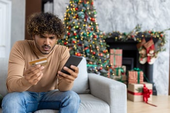 An upset man looks at his phone and credit card in front of a Christmas tree
