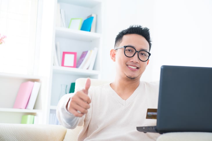 A young man smiles and gives a thumbs up from behind his laptop