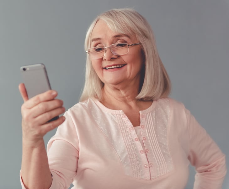 An older woman in glasses smiles while holding a smartphone