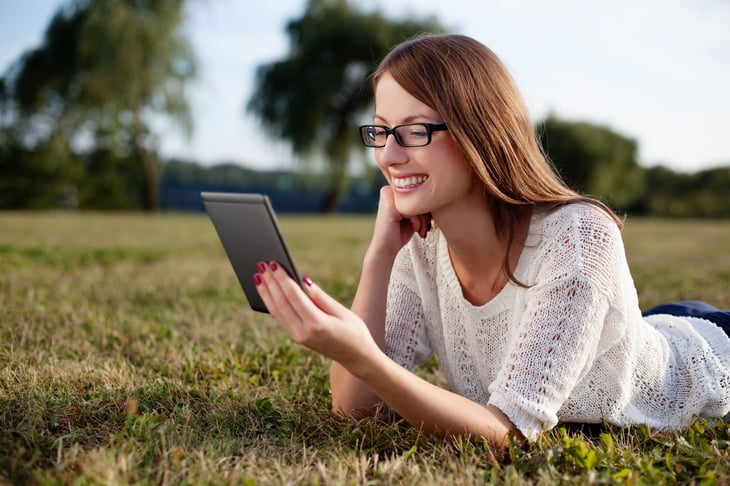 A woman smiles while enjoying reading an e-book on an e-reader and lying on the grass outside