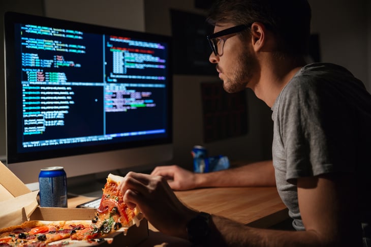 A computer programmer eats pizza while working at his desk