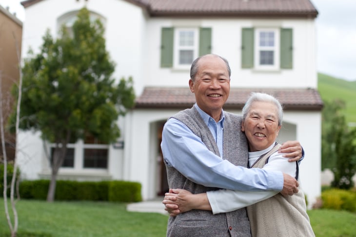 An older couple stands in the front yard of their house
