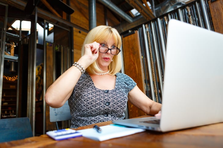 A woman in glasses looks at a laptop computer screen in surprise