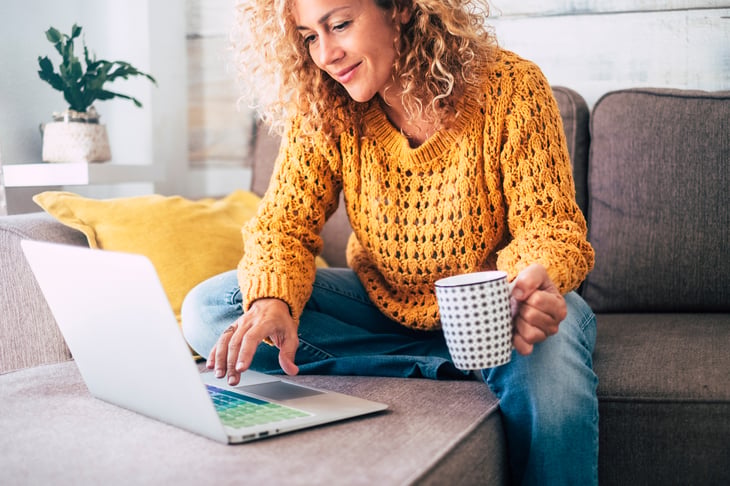 A woman drinks from a coffee mug and types on a laptop computer keyboard while sitting on her living room sofa at home