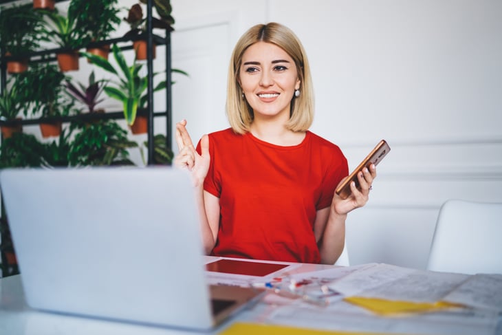 A woman holds her phone in one hand and crosses her fingers with the other while working at her laptop computer