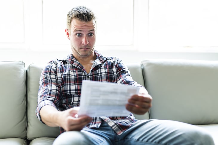 A man reads a bill in shock while sitting on his couch at home