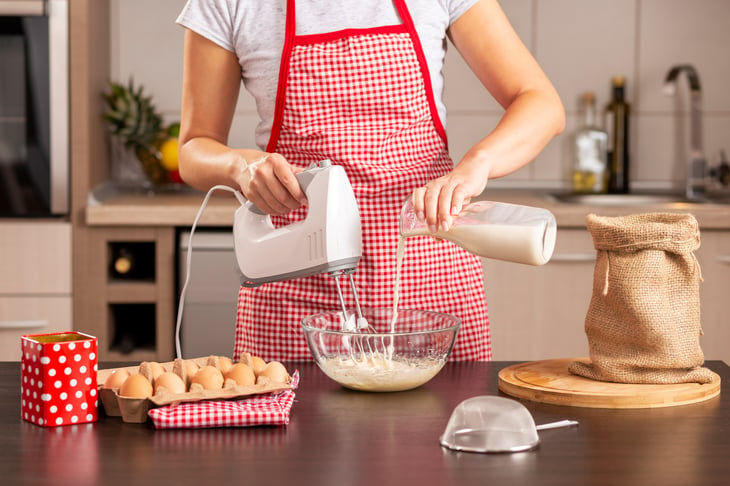 A woman in an apron uses a handheld mixer while baking in her home kitchen