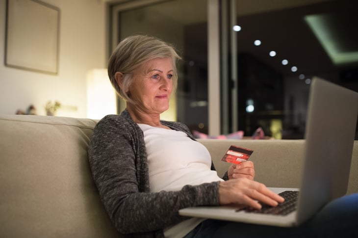 A senior shops online from her living room sofa with a laptop computer and credit card