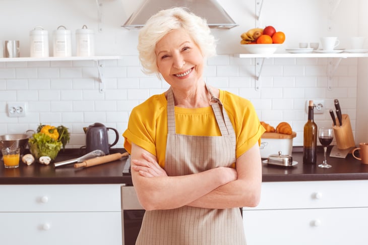 An older woman in an apron smiles in her kitchen