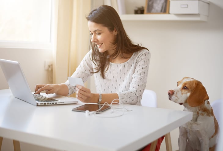 A woman works on a laptop computer from home next to her dog