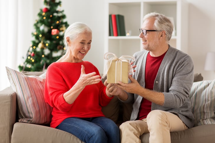 A senior husband presents a holiday gift to his wife in front of a Christmas tree