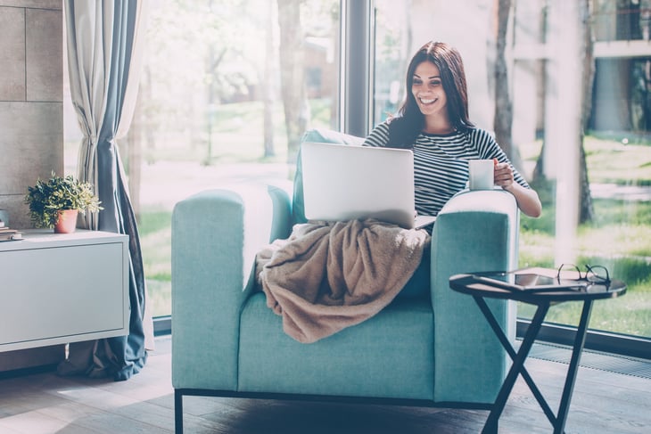 A woman works from home on a laptop computer while drinking from a coffee mug and sitting under a blanket on a living room chair