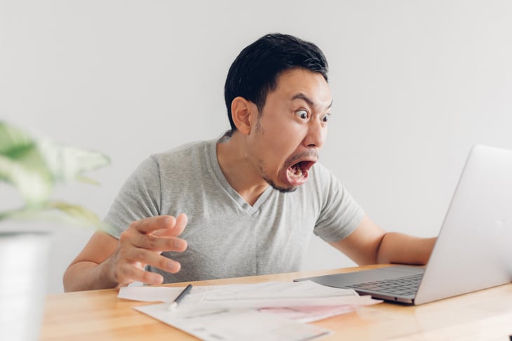 A young Asian man at his laptop computer is surprised and shocked by a bill
