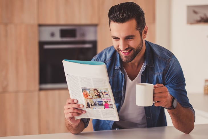A man reads a magazine in his kitchen while drinking from a mug