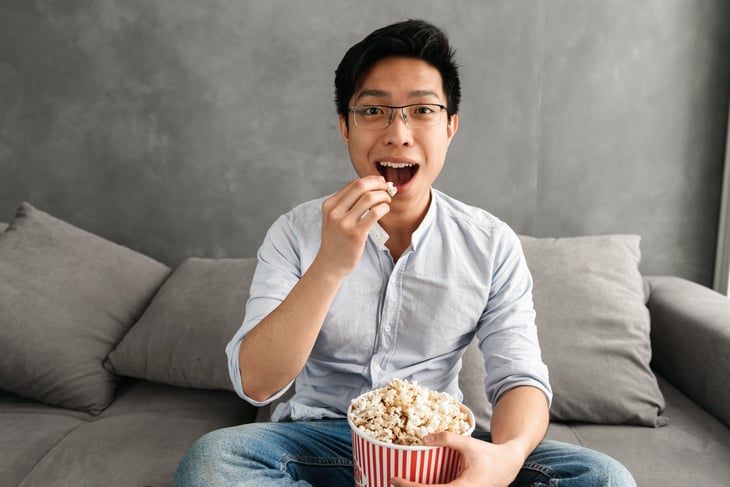 A man in glasses eats popcorn on his couch while watching TV