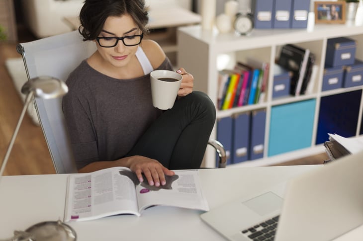 A woman in glasses reads a magazine at her desk while drinking a mug of coffee