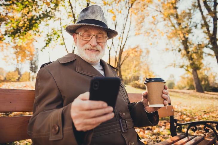 Happy senior man sipping coffee in the park on an autumn day and checking his smartphone