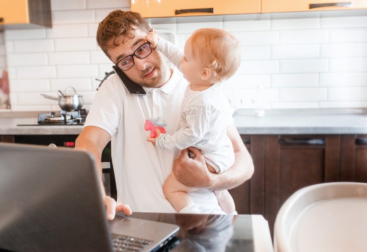 Young father holding a baby and trying to get work done while on the phone and laptop