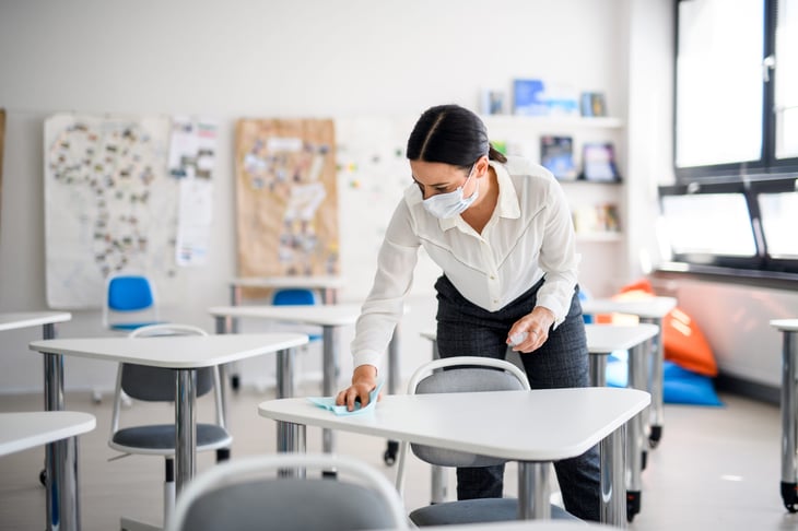 Teacher in a face mask disinfecting student desks