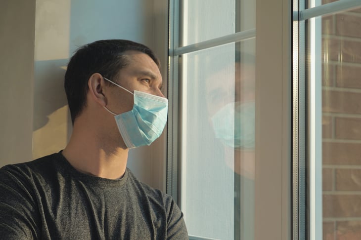 Worried man in a mask looking out a window while self-isolating