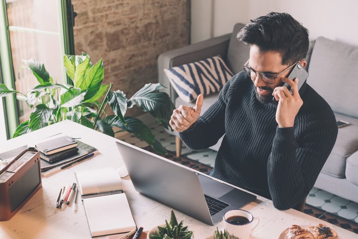 Man on his phone, working remotely from his home office with a laptop, notebook, and coffee