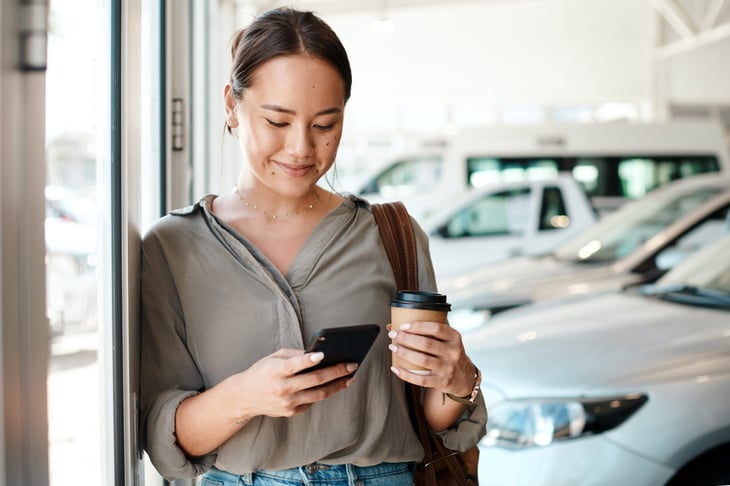 Woman with coffee using her smartphone at a car dealership to find the best deal.