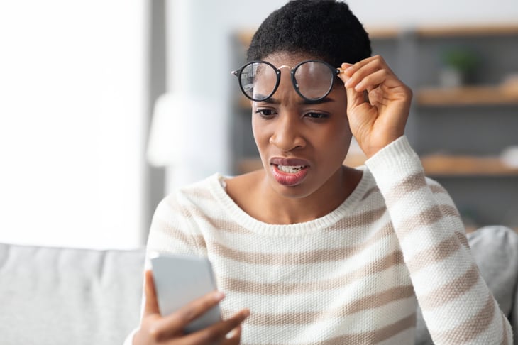 Upset woman raising her eyeglasses in surprise while looking at her phone
