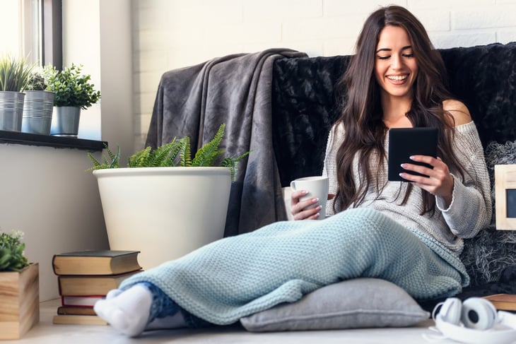 Cozy happy woman reading an ereader or Kindle with a blanket and cocoa or coffee