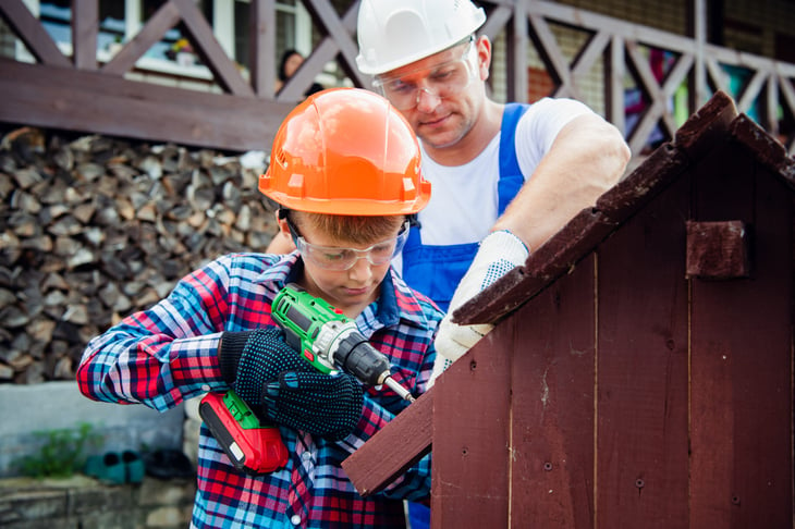 Father and son building a dog house with drill and wearing construction helmets