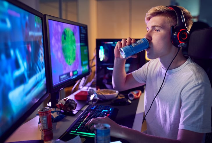 Young man gamer playing video games on computer and drinking energy drink