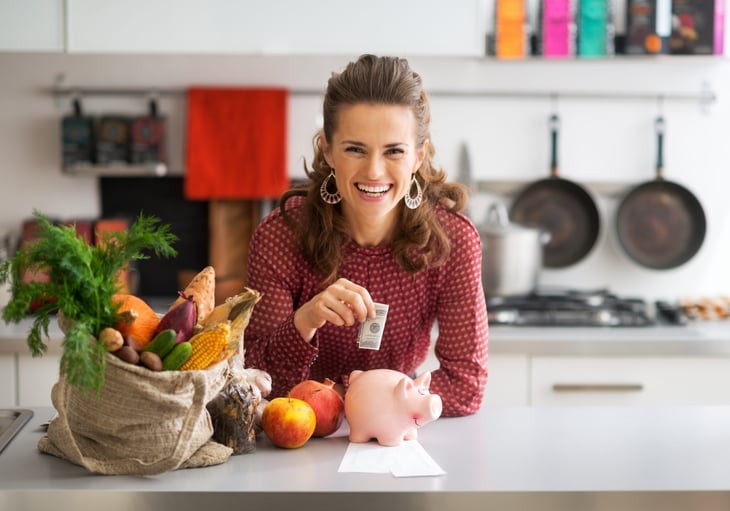 happy woman in kitchen putting money in piggy bank